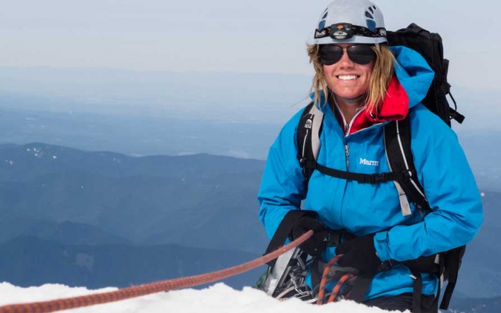 A person wearing mountaineering gear is secured by a rope as they stand on a snowy cliff and smile at the camera. There is a vast mountainous landscape in the background.